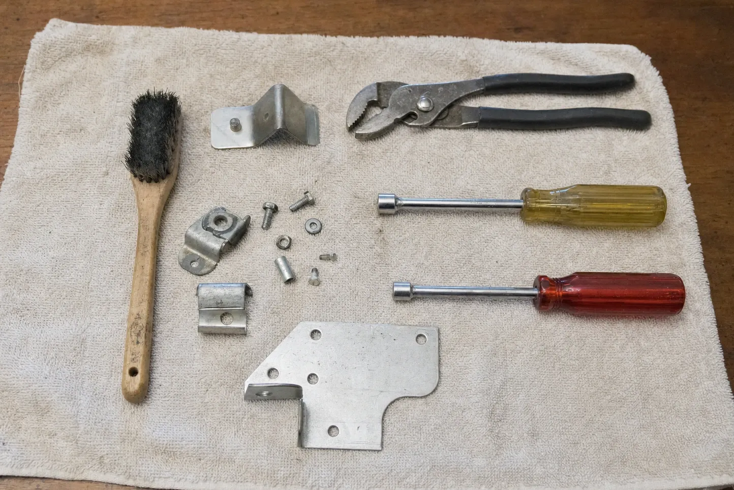 Wire brush, hinge plate and brackets, screws and a ferrule set out on a cloth next to slip-joint pliers and two nut drivers