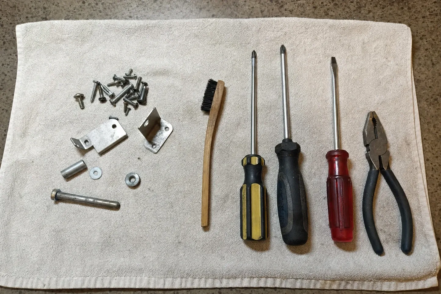 Screws, hinge brackets, a roller pin and washers heaped on a cloth beside a wire brush, two Phillips drivers and pliers