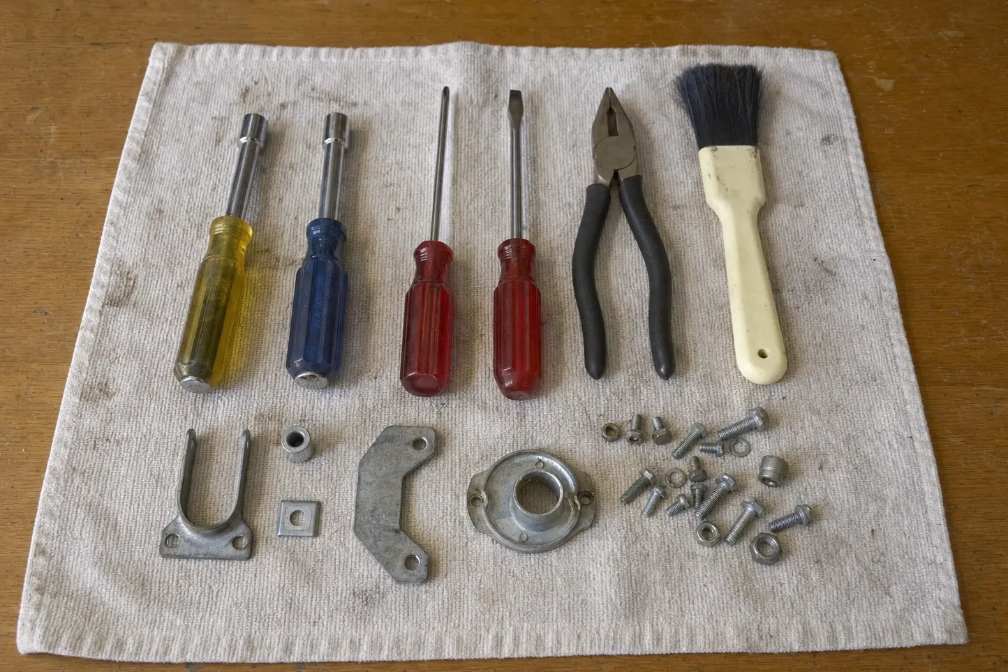 Two nut drivers, two red-handled screwdrivers, pliers and a bristle brush on a ribbed towel above a forked bracket, a round flange collar, a curved plate and loose bolts